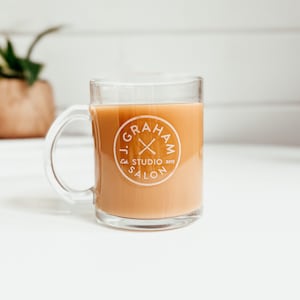 May include: A clear glass mug with a brown beverage inside. The mug has a white logo that says "J. Graham Studio Salon Est. 2013" with a pair of crossed scissors inside a circle.
