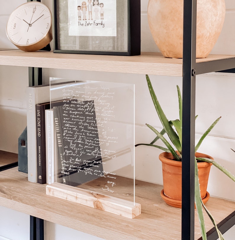 K&ouml;nnte beinhalten: Eine klare Acrylplatte mit einer handgeschriebenen Nachricht auf einem Holzregal. Die Nachricht ist ein herzlicher Brief, der mit wei&szlig;er Tinte geschrieben wurde. Die Platte steht aufrecht auf einem Holzsockel. Auf dem Regal stehen B&uuml;cher und eine Topfpflanze.