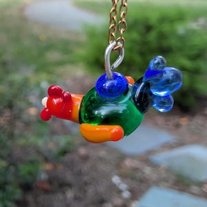 Puede incluir: Un colgante de avión de cristal colorido suspendido de una cadena dorada. El avión en miniatura presenta un cuerpo verde, alas naranjas, cola roja y detalles azules. El colgante mide aproximadamente 2.5 cm de largo, perfecto para un collar.