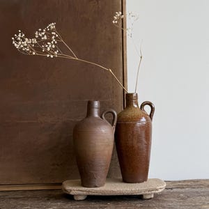 Peut inclure: Deux vases en céramique marron avec des anses reposent sur une petite plateforme rectangulaire beige. Les vases ont un aspect rustique et vieilli. Une brindille de fleurs blanches séchées dépasse du vase de droite. Le fond présente un panneau marron et un mur blanc.