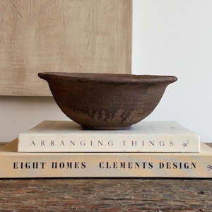 May include: A dark brown, textured ceramic bowl with a wide, slightly flared rim. The bowl is resting on top of two stacked hardcover books titled Arranging Things and Eight Homes Clements Design, which feature neutral-toned covers.