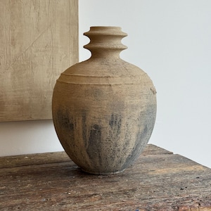 May include: A round, textured ceramic vase with a narrow, tiered neck. The surface features a weathered, earthy tan finish that transitions into a dark, charcoal-grey mottled pattern towards the base, creating a rustic and aged appearance.