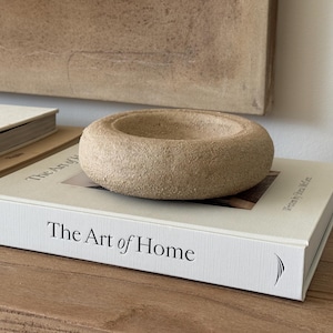 Könnte beinhalten: Eine beige, kreisförmige, steinartige Schale liegt auf einem Buch mit dem Titel "The Art of Home". Das Buch ist weiß mit schwarzem Text. Weitere Bücher sind im Hintergrund zu sehen. Die Szene spielt auf einer Holzoberfläche.
