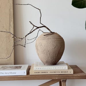 Puede incluir: Un jarrón beige texturizado con un arreglo de ramas, apoyado sobre una pila de libros. Los libros se titulan "The Art of Home" y "Arranging Things". El jarrón está sobre una mesa de madera, con una pared de tonos neutros y una planta en el fondo.