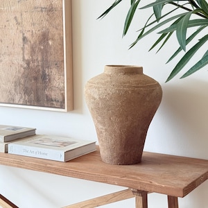 Puede incluir: Un jarrón beige grande y texturizado, con una base ancha y un cuello estrecho, está sobre una mesa de madera. Dos libros y una obra de arte enmarcada también están sobre la mesa. Hojas verdes en la esquina superior derecha.