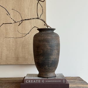 Könnte beinhalten: Eine dunkelbraune, verwitterte, zylindrische Vase mit einem ausgestellten Rand und einer texturierten, matten Oberfläche. Ein einzelner, kahler, knorriger Zweig ragt aus der Öffnung. Die Vase steht auf zwei gestapelten Büchern, das obere trägt den Titel CREATE AT HOME WITH OLD & NEW.
