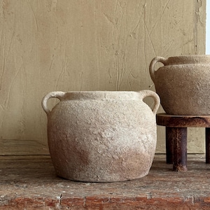 May include: Two textured, beige ceramic vases with rounded bodies and loop handles. One sits on a wooden surface, the other on a small, dark wooden stand. The background is a neutral, textured wall. The vases are suitable for home decor.