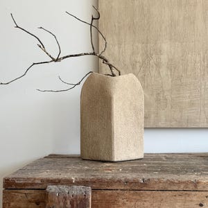 May include: A beige, textured, square-shaped vase with a matte finish. The vase has a wide, open top and features a single, thin, dark brown, leafless branch extending from the opening, creating a stark contrast against the light-coloured vessel.