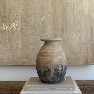 May include: A textured, round, beige and dark grey ceramic vase with a wide neck, containing a delicate sprig of dried baby's breath. The vase rests on two stacked books, one titled The Space That Keeps You by Jeremiah Brent and the other Pablo Picasso.