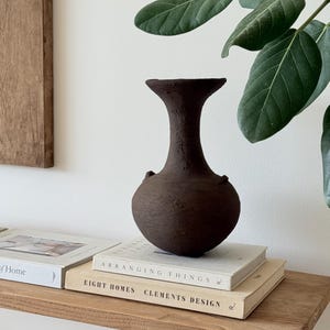 Peut inclure: Un vase en argile texturé brun foncé avec un corps arrondi, un col étroit et un bord évasé. Le vase présente deux petites anses subtiles sur les côtés et est posé sur deux livres empilés intitulés Arranging Things et Eight Homes.