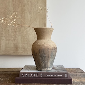 Puede incluir: Un jarrón de cerámica texturizado en tonos beige y gris oscuro con borde acampanado y base cónica. El jarrón contiene una delicada rama de flores blancas secas y descansa sobre dos libros apilados, uno titulado CREATE AT HOME WITH OLD & NEW.