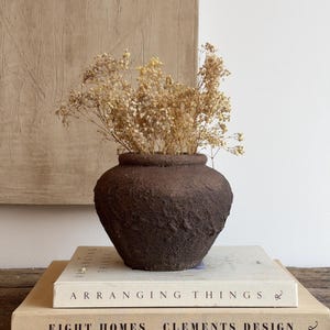 May include: A dark brown, textured, round ceramic vase containing a bouquet of dried, light-colored baby's breath flowers. The vase sits atop two stacked hardcover books titled Arranging Things and Eight Homes Clements Design.