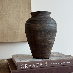 Puede incluir: Un jarrón de arcilla de color marrón oscuro con textura, cuerpo ancho y redondeado y cuello estrecho. El jarrón tiene una superficie rugosa y desgastada y se apoya sobre dos libros apilados, uno titulado CREATE AT HOME WITH OLD & NEW y el otro JAKE ARNOLD.