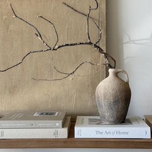 May include: A textured ceramic vase in shades of brown and grey, with a handle, sits on a shelf with books, including "The Art of Home." A dried branch extends from the vase, against a textured beige backdrop. The scene evokes a sense of rustic elegance.