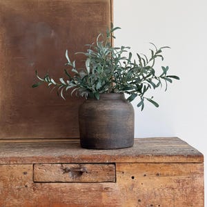 May include: A dark brown, textured ceramic vase filled with green leafy branches. The vase sits on a weathered wooden table with a small drawer. A framed piece of art with a brown background is behind the vase.