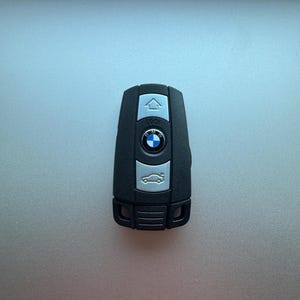 May include: Black car key with silver buttons and a BMW logo. The key has a rectangular shape with rounded edges. The buttons include lock, unlock, and a car icon.