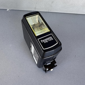 May include: Black Panasonic PE-14S camera flash with a rectangular shape. The flash has a clear, yellow-tinted lens and a silver-colored mounting bracket. The brand name and model number are printed in white on the front.