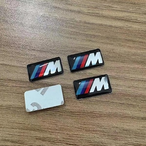Op de afbeelding: Vier rechthoekige auto-emblemen met de letter "M" in het wit en een ontwerp met blauwe, rode en paarse strepen. Eén embleem heeft een witte zelfklevende achterkant. De emblemen liggen op een houten oppervlak.