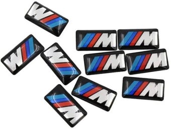 Puede incluir: Un conjunto de emblemas rectangulares negros para automóviles con las letras "M" y "W" en blanco. Cada emblema presenta un diseño de rayas rojas, azules y celeste. Los emblemas son brillantes y parecen ser de plástico.