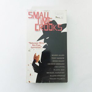 May include: A VHS tape for the movie "Small Time Crooks" with a white background. The title is in red letters. A black silhouette of a man in a hat is holding a cookie. The cast and a quote are also visible.