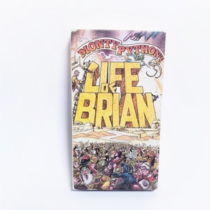 May include: A vintage paperback book titled "Monty Python's Life of Brian." The cover features a large yellow title with a crowd of people at the bottom and illustrations of characters and scenes from the film.
