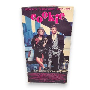 May include: A vintage VHS tape for the movie "Cookie." The cover features Peter Falk, Dianne Wiest, and Emily Lloyd. The title "Cookie" is in pink bubble letters. The background shows a city skyline and two people standing near a car. The tagline reads "A delicious new comedy."