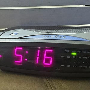 May include: A black digital clock with a pink display showing the time 5:16. The clock has a radio and alarm function and a snooze button. The clock also has a battery low indicator and a green light indicating the time is set.