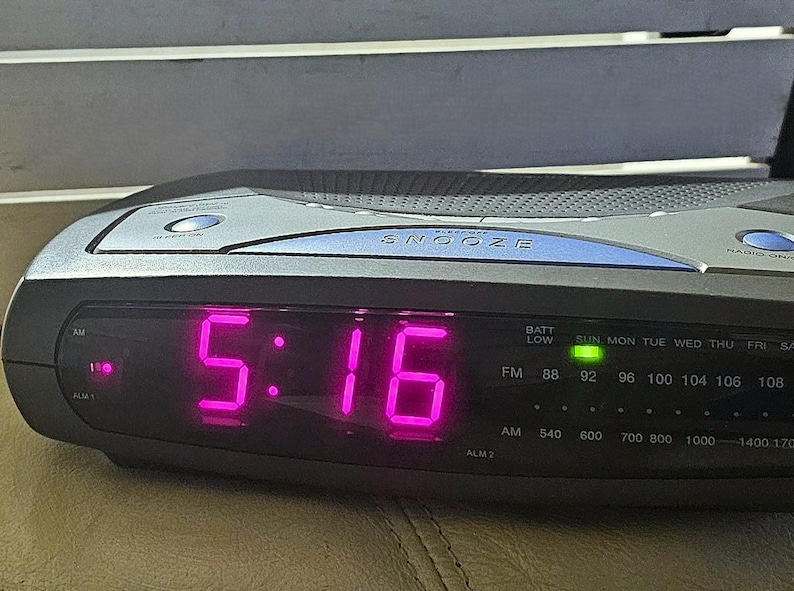 May include: A black digital clock with a pink display showing the time 5:16. The clock has a radio and alarm function and a snooze button. The clock also has a battery low indicator and a green light indicating the time is set.