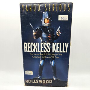 May include: A VHS video cassette cover with a blue background and the title "Reckless Kelly: The Incredible Untold Story of the Greatest Outlaw of All Time."  The cover features a man in a silver helmet and armor holding two guns.