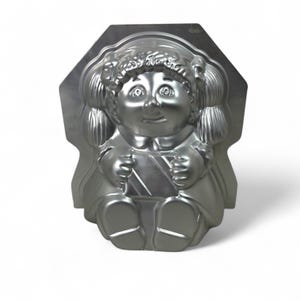 May include: A silver-colored metal baking mold in the shape of a doll. The doll has a smiling face, pigtails, and is wearing a dress. The mold is designed with a detailed, three-dimensional appearance, perfect for creating shaped cakes or desserts.