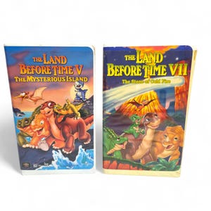 May include: Two VHS tapes of "The Land Before Time" animated movies. The first tape is titled "The Land Before Time V: The Mysterious Island" and the second tape is titled "The Land Before Time VII: The Stone of Cold Fire". The tapes feature colorful illustrations of dinosaurs.