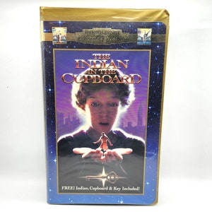 May include: A VHS tape case for the movie "The Indian in the Cupboard". The case is gold with a blue background and features a picture of the main character holding a small figure in his hand. The text "FREE! Indian, Cupboard & Key Included!" is printed on the bottom of the case.