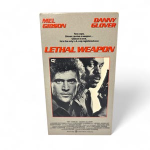 May include: A vintage VHS tape for the movie "Lethal Weapon" featuring Mel Gibson and Danny Glover. The cover has a gray background with the title in orange and images of the actors. The tape is in a vertical format.