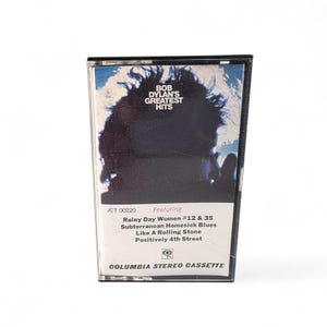 May include: A vintage cassette tape of Bob Dylan's Greatest Hits. The album cover features a dark silhouette with the text "BOB DYLAN'S GREATEST HITS". The tracklist includes "Rainy Day Women #12 & 35" and "Like A Rolling Stone". The cassette is labeled "COLUMBIA STEREO CASSETTE".