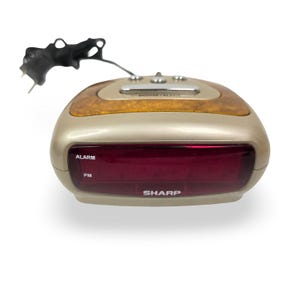 May include: A gold-colored Sharp digital alarm clock with a wood-grain top and a red digital display. The clock has a snooze/alarm button and a black antenna. The word "SHARP" is printed on the front.