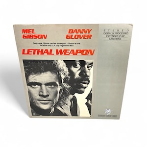 May include: A LaserDisc movie cover for "Lethal Weapon" featuring Mel Gibson and Danny Glover. The cover has a gray and white design with the movie title in red. The Warner Bros. logo is visible.