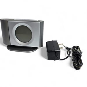May include: A silver and gray digital alarm clock with a circular display. The clock has a rectangular shape and sits on a small base. A black power adapter with a cord is next to the clock.