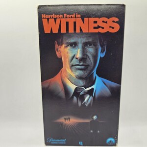 May include: A VHS video cassette cover featuring Harrison Ford in the movie "Witness". The cover is red, orange, and blue with a dark background. The title "Witness" is in large, bold, red letters. The image shows Harrison Ford looking directly at the viewer.