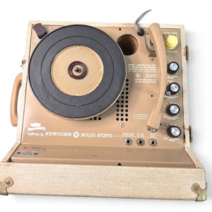 May include: A portable Newcomb Solid State record player in a beige textured case. The device features a black turntable platter, a beige tone arm, and a control panel with four black knobs, a yellow knob, and three input jacks. Text includes Newcomb Solid State.