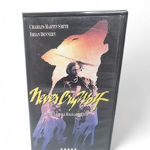 May include: A VHS tape for the film "Never Cry Wolf" featuring Charles Martin Smith and Brian Dennehy. The cover art depicts a man in silhouette against a sunset with a wolf. The film title is in yellow script.