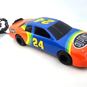 May include: A toy race car telephone in blue, orange, and yellow with the number 24 on the roof and side. The phone has a black cord and is branded with "DuPont".