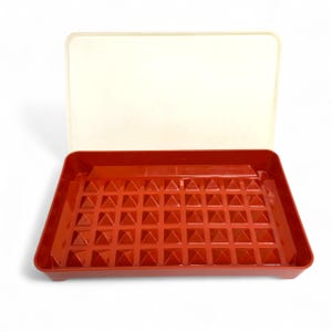 May include: A red plastic tray with a grid of pyramid shapes, paired with a cream-colored lid. The tray is rectangular and has a raised edge. The lid is also rectangular and sits on top of the tray.