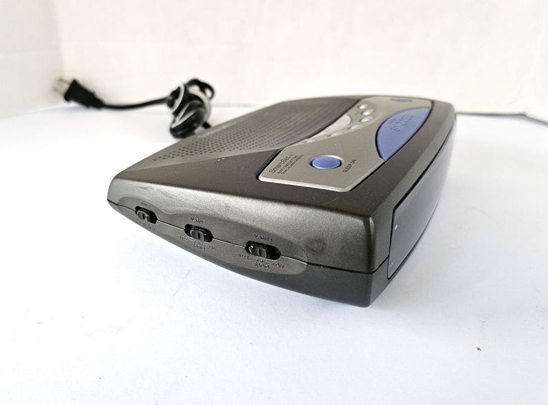 May include: A black and silver clock radio with a blue button and a small LCD screen. The radio has several buttons on the side, including "Sample", "Mute", and "Aux In".