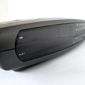 May include: A black digital clock radio with a large display screen. The clock radio has buttons for AM, ALM 1, and other functions.