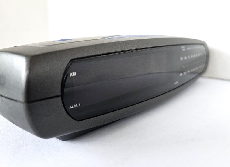May include: A black digital clock radio with a large display screen. The clock radio has buttons for AM, ALM 1, and other functions.