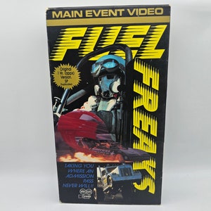 May include: A VHS video cassette with a black background and yellow text that reads "Main Event Video" and "Fuel Freaks". The image shows a driver in a race car with flames coming from the exhaust.