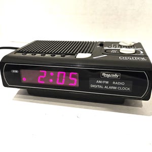 May include: Black digital alarm clock with a pink display showing 2:05 PM. The clock has a built-in AM/FM radio and is labeled "Rhapsody Digital Alarm Clock".
