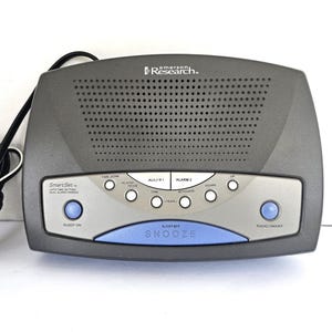May include: A black Emerson Research SmartSet clock radio with a blue sleep/snooze button and a silver face. The clock radio has two alarm settings, a radio tuner, and a sleep timer.