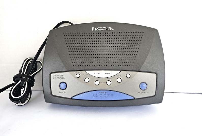 May include: A black Emerson Research SmartSet clock radio with a blue sleep/snooze button and a silver face. The clock radio has two alarm settings, a radio tuner, and a sleep timer.