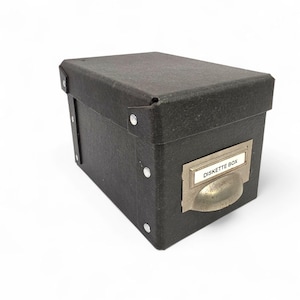 May include: A black rectangular storage box with a lid. The box has a metal label holder with the words "DISKETTE BOX" printed on a white label. The box is made of a textured material and has metal rivets.